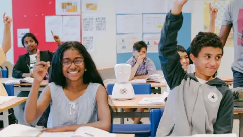 Pupils sit at desks in a classroom, many with their hands up and smiling, while a white robot is placed on one of the desks 