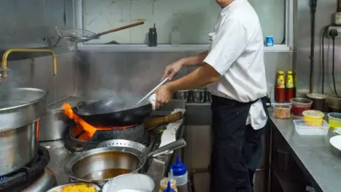 A stock image showing a man wearing black trousers, a white chef's shirt and a black apron. He is using a spatula to stir ingredients in a large wok, which is resting on an open flame beside other pots and pans. He is standing in a commercial kitchen between metal counters with various condiments in plastic containers. 