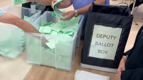 Two transparent plastic containers, stuffed with green voting slips. A number of hands are rummaging around in the containers. To the right of the image is a dark container which is labelled 'Deputy Ballot Box'.