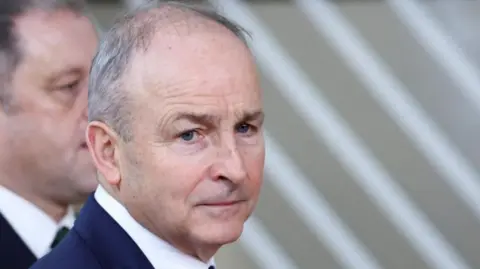  Micheál Martin is looking at the camera but his head is turned to the side. He has short grey hair.