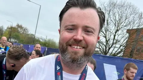 James Derrig A man smiles at the camera. He is wearing a white T shirt and a medal around his neck. 