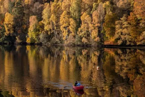 Sophie Coleman Trees with orange and green leaves reflecting onto Loch Faskally. There is two people in a kayak in the foreground.