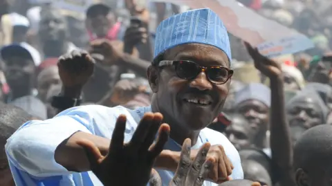 At a packed rally, supporters clamour to touch Buhari, who stands above the crowd, smiling. He wears a blue top and blue hat.