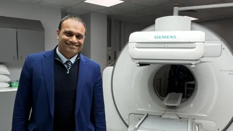 Dr Randeep Kulshrestha stands in front of a medical scanner. He wears a blue suit, with a blue shirt, striped tie and navy quarter zip jumper. Behind him is a large circular white scanner. 