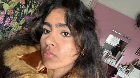 A young woman with long dark wavy hair and a fringe looks directly into the camera in a selfie taken from a slightly elevated angle. She wears a brown and tan faux fur coat with a satin collar and has dark eye makeup on. Behind her is a pink-walled bedroom with a floral feature wall, a white chest of drawers, a small mirror and a desk fan visible in the background.