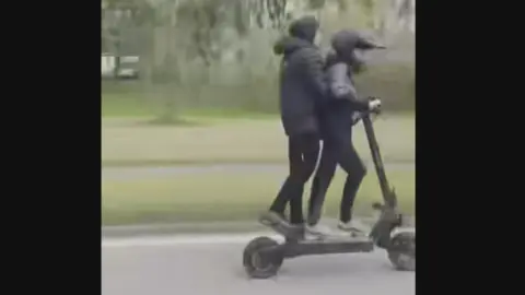 Two youths on an e-scooter on a public road with trees behind.