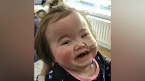 Family handout Another image of Lila - this time she has her hair in a bobble on top of her head and is smiling. She's wearing a dark top with a pink border at the neckline. A tube is coming out of one of her nostrils.