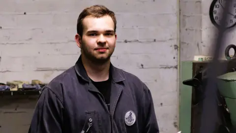 Ernest Wright Elliott Nurcombe in a blue boiler suit stands in the Ernest Wright workshop