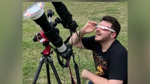 Josh Dury Josh Dury wearing eclipse glasses and looking up towards the sky.
He has a telescopic camera next to him and is kneeling on grass. 