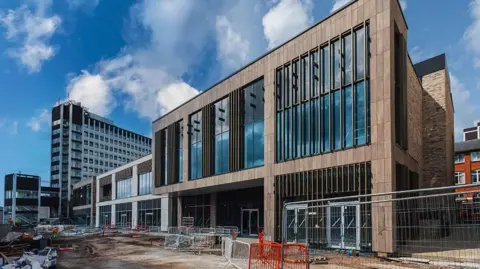 The Animate building, under a blue cloudy sky, showing a large rectangular two-storey building with brindle brick and huge glass windows. There is a tall tower block to the left and construction barriers can been seen to the front where the road and pavement are unfinished.