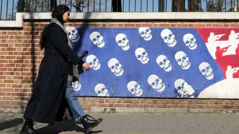 A woman in black in Iran walks near a mural that depicts the US flag with skulls instead of stars