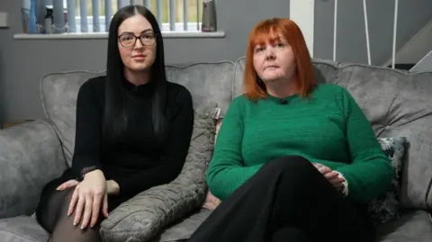 Laura and Elisha sit on a grey sofa and look at the camera. Laura has red hair and is wearing a bright green jumper and black trousers, while Elisha has long dark hair, glasses , and is wearing a black dress.