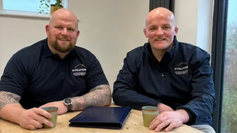 Supplied Jake, pictured on the left, sits next to his brother Josh at a wooden table. Jake wears a navy blue polo shirt with the Evolution Development South West logo on the right breast. He is holding a green coffee cup in one hand. Josh is on the right, also wearing a company jacket and holding a green coffee cup in his hand. They both smile at the camera.