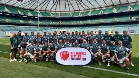 A rugby team is sitting on a rugby field in their kit. Some have their arms around each other. They are holding a sign with the RFU injured players foundation logo on it.