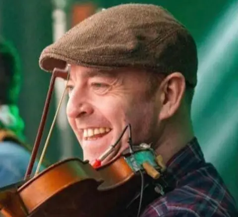Facebook A man smiling and wearing a brown flat cap and red, white and dark blue flannel top. He has a violin under his chin