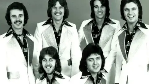 Pacemaker A black and white image of the band members. They all have dark long hair with some also having moustaches. They are wearing blazers with patterned shirts.