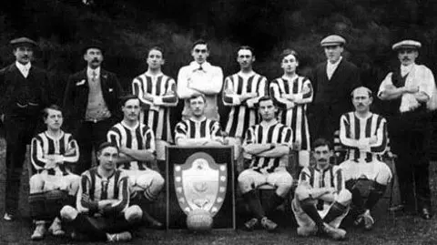 RH Clark A black-and-white team photo from when the club won their first trophy in the 1911-12 season. Fifteen men pose for the photo around a large framed shield. Some of the men are dressed in player kit of a black and white striped shirt, shorts and long socks. Four of the men are wearing flat caps. 