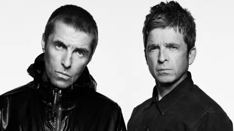 PA Media Black and white image of Liam and Noel Gallagher. Both are looking at the camera with serious faces. Liam is on the left and is wearing a shiny jacket. He has short dark hair. Noel, on the right, is wearing a denim shirt. He has short spiky hair.