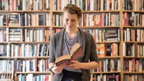 A woman standing in front of a wall full of laden bookshelves. She is wearing a long grey cardigan and blue striped top, and looking at an open book in her hands.