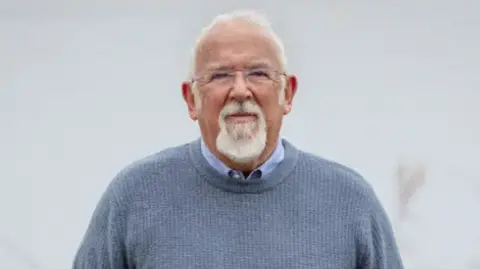 A man wearing a grey blue jumper and blue shirt stands looking at the camera. He has a short white beard and short white hair. He is wearing glasses.