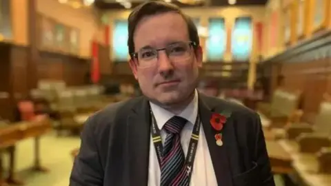 Lawrie Hooper, who is standing in an empty House of Keys chamber. He is wearing glass and has dark hair and a close-cropped beard. He is wearing a grey suit and a striped tie. 