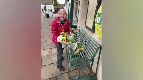 Anne Cradock is holding a bunch of flowers and is standing in front of a green metal bench which also has two bunches of flowers placed on it. Anne has short white/grey hair and is smiling broadly at the camera.