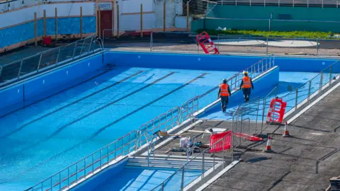 Marcin Jedrysiak A wide view of one end of the pool, with a small amount of water in it, with two workmen in orange hi-vis vests walking alongside