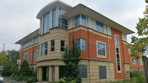 Google The front view of a large multi-storey building. There are trees and greenery surrounding it. 