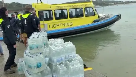 Above and Below Imagery Bottled water is piled up on Bryher's quayside. South West Water set up the bottled water station while a faulty pipe was repaired on the island. A yellow boat is in the background. Several people are stood on the quayside.