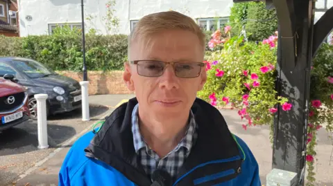Sam Read/BBC John Baker with short white hair, wearing glasses and a blue anorak over a blue and white checked shirt. He is standing in front of pink roses to the right and a car park to the left. There is an office building behind.