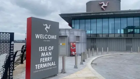 A welcome to the Isle of Man ferry terminal freestanding sign stands to the left and in front of the glass panelled building, with a triskelion symbol above it.