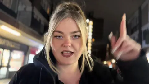 Still from a Morgan da Savage rap video shows her mid-flow, staring down the barrel of the camera as she points her finger at the sky. She's standing in the middle of a row of shops at night, with a tower block occupying the background of the shot.