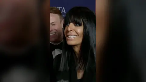 Claudia Winkleman smiling. She has a man stood behind her and a blue promotional Bafta backdrop.