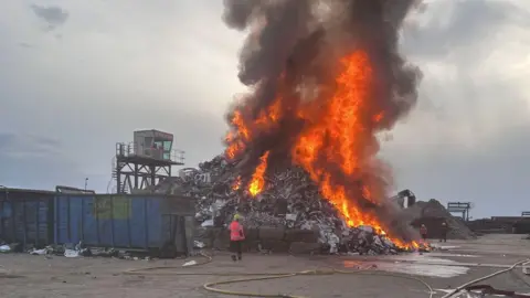 A photo taken outside during daylight hours. A huge heap of metal and plastic is stacked up and on fire. IT looks like there are washing machines among the waste. Three firefighters stands around the outside, and long yellow hoses lie on the floor.