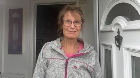 A woman with brown hair tied up. She has glasses and is standing in a doorway with the white door open. She is wearing a grey and pink zip hoodie.