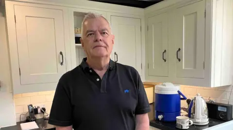 A man in a black polo top standing in a kitchen with white cupboard doors and a black granite work surface.