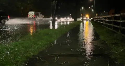 BBC Water spray is visible near two vehicles on a road on the left of the photo in the dark. Water is also visible on a pavement on the right. Other vehicles are in the middle of the image with their lights on.
