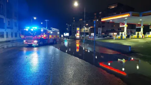 Mark Merely A fire engine with its blue lights on is parked on a road which is partially flooded next to a petrol station