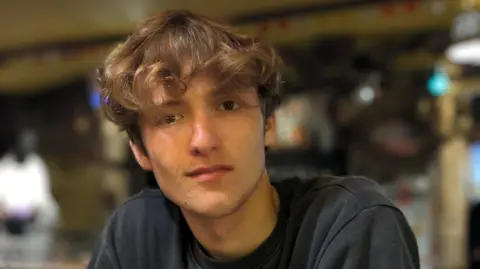 Arlo looks directly at the camera, with a small smile. He has brown eyes and light brown/blonde tinted curly hair framing his face. Arlo wears a black round neck sweatshirt and the background behind him is out of focus.