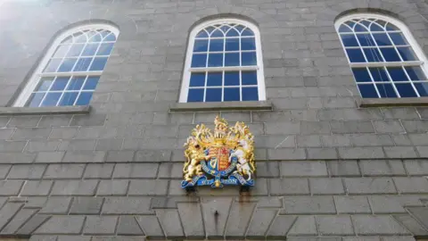 Grey brick building with the royal coat of arms on the wall and large windows with curved tops.