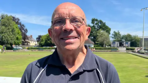 Mark looking into the camera and smiling. He has a bald head and is wearing a navy blue short-sleeved sports shirt. He is standing in front of a bowling green, which is green and grassy. The sky is bright blue with a few clouds, and it is very sunny.