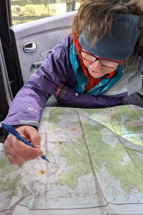 Lorraine McCall A woman in a purple jacket is studying a map planning a walking route. She is wearing glasses and is holding a pencil to annotate the map.