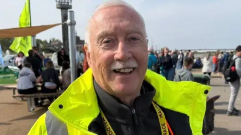 A man with grey hair and a grey moustache smiles at the camera. He wears a bright yellow hi-vis coat with a black jumper underneath and a yellow lanyard around his neck. People can be seen milling about behind at an event.