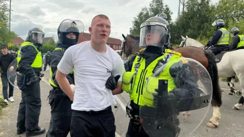 BBC/Oli Constable Gray is arrested by two officers who are wearing riot shields and helmets. A mounted officer can be seen in the background together with several other officers on foot. Gray is wearing a white t-shirt and black trousers. He has blond, shaven hair.