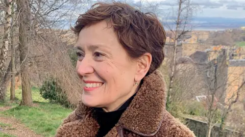 Carys Davies is smiling as she looks to the left of the camera. She has short brown hair and is wearing a black top with a brown fleece jacket. She is in the countryside, with trees behind her.