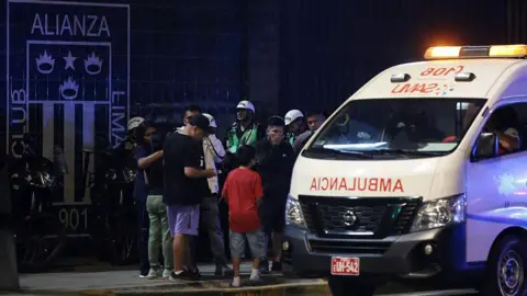 Getty Images Crowds of people gathering near an ambulance at one of the entrances of the Alejandro Villanueva Stadium in Lima, Peru. It's night time and some of the fans are hugging, others looked shocked.