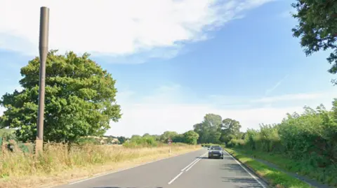 A Google Maps street view image of a road which is flanked by trees and grass verges. A car is travelling towards the camera.