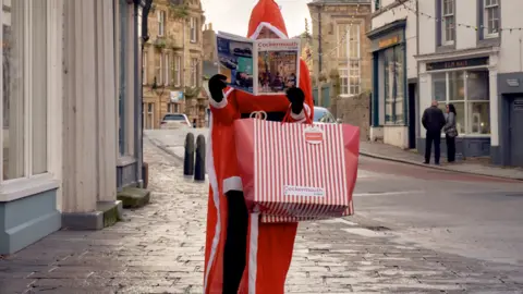 A woman dressed in a red Santa suit, holding a newspaper and a shopping bag, standing on a cobbled street. 