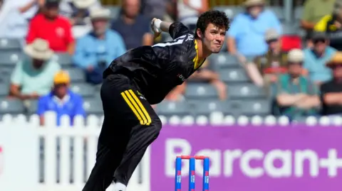 Ollie Price bends forwards after bowling a ball during a match for Gloucestershire. He is wearing black team kit