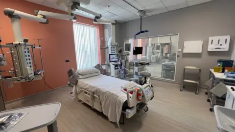 The picture shows a hospital room set up to deliver critical care. There is a bed in the middle of the room that is surrounded by medical equipment. The room is large and spacious. One of the walls is grey, while the other is burnt orange.